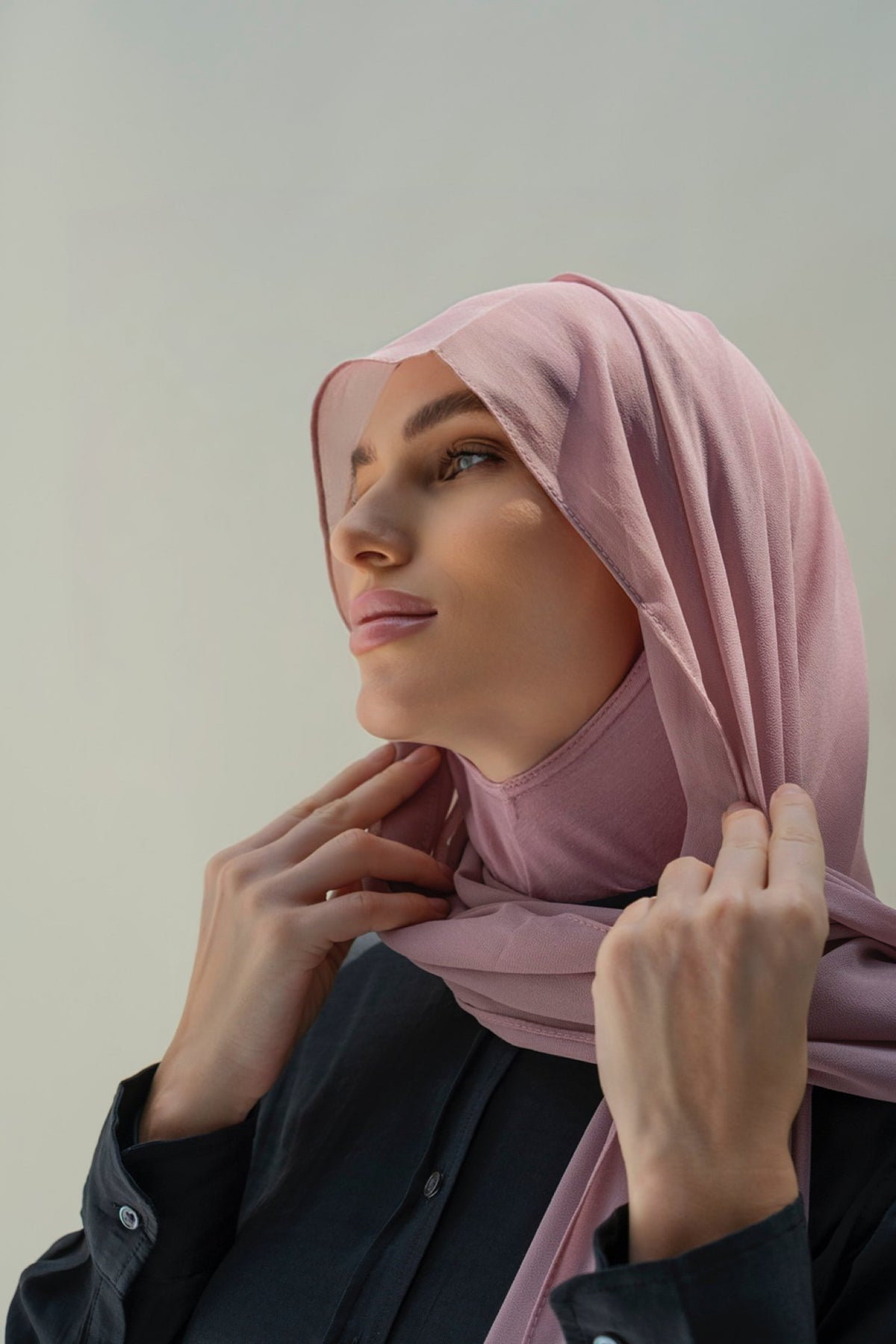 A woman wearing a dusty pink hijab and a black button-up shirt, posing against a neutral light gray background.