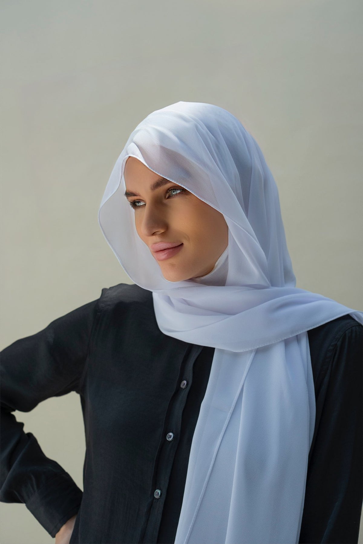 A woman wearing a light blue semi-sheer hijab and a black button-up shirt, photographed against a neutral light gray background.