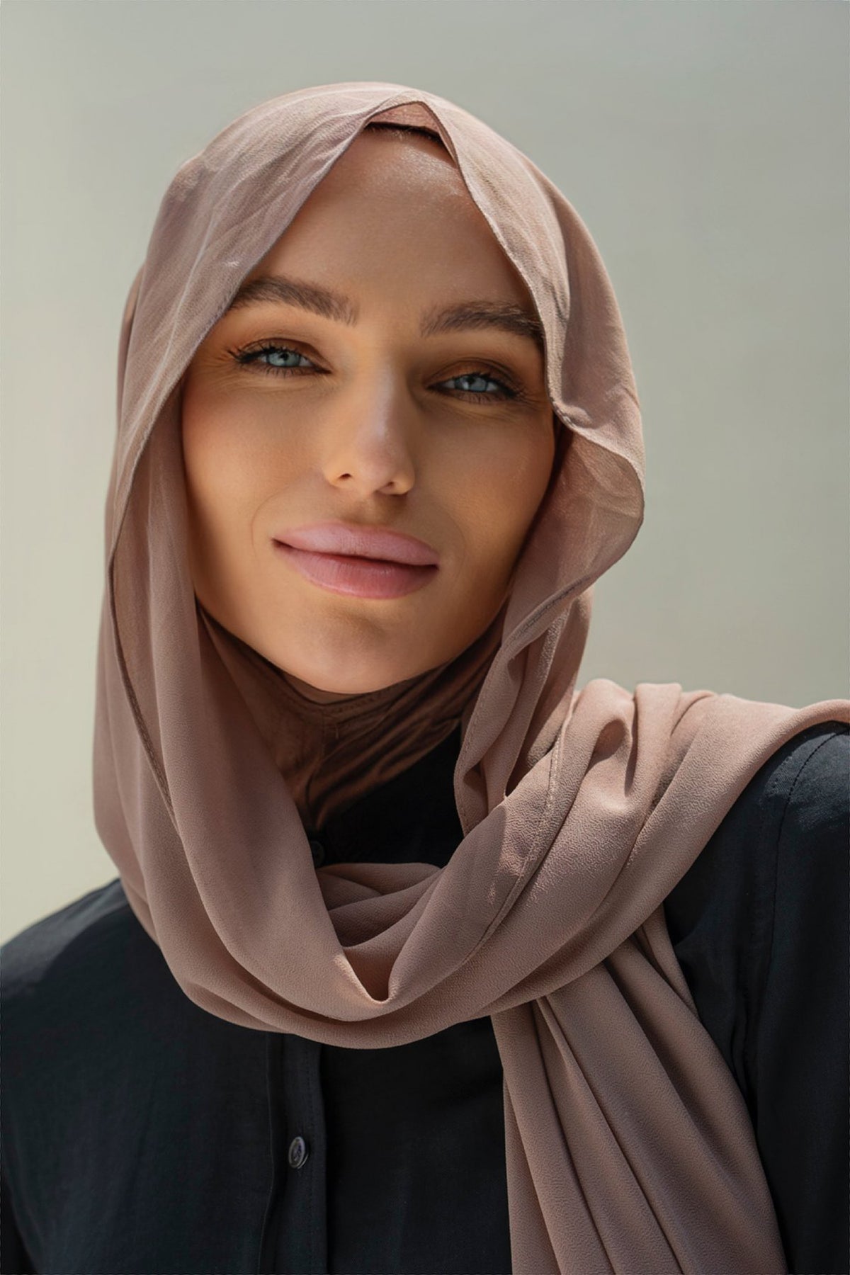 A close-up portrait of a woman wearing a draped dusty rose hijab and a black top, set against a soft, neutral gray background.