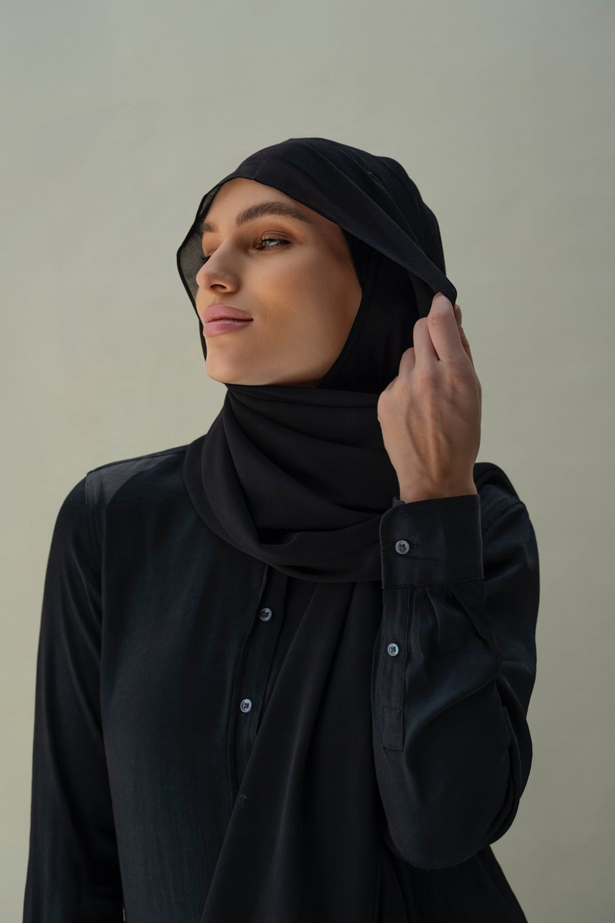 A woman wearing a black hijab and a black button-front top, posed against a plain light gray background.