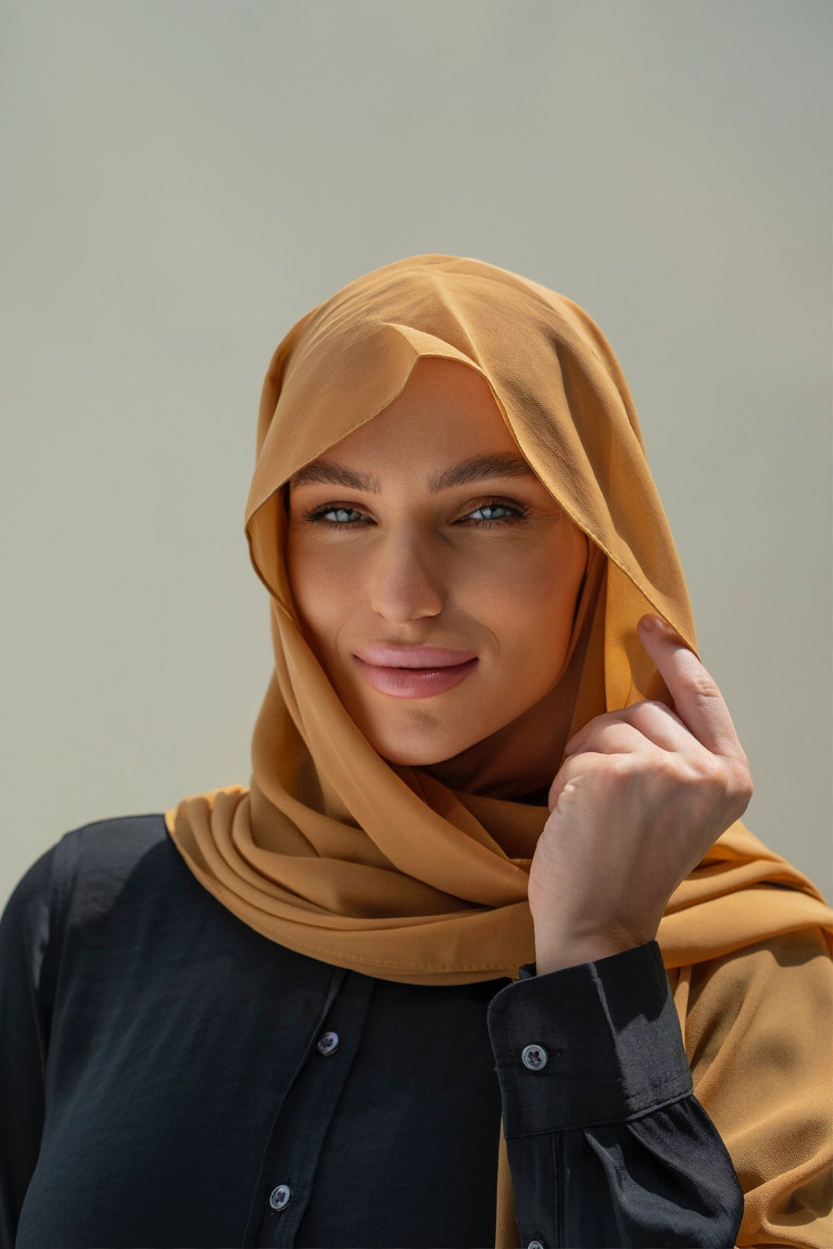 A woman wearing a mustard yellow hijab and a black button-up shirt, posing against a neutral light gray background.