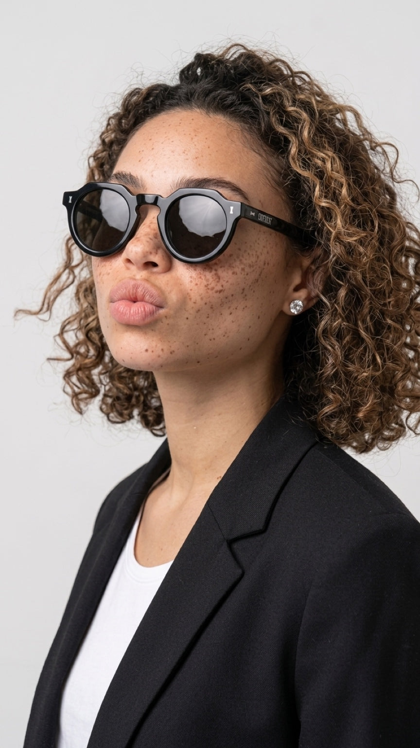 A woman with curly brown hair wearing round black CONFILVARO Black Round Sunglasses
with dark lenses, a black blazer, and a white top, photographed against a light gray studio background.