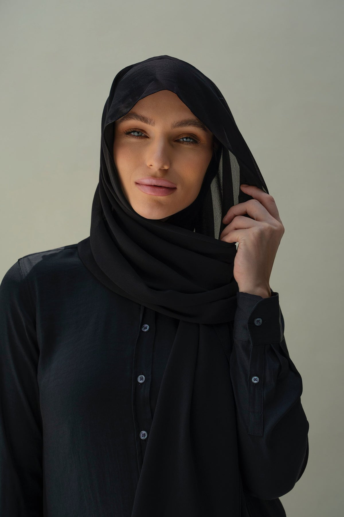 A woman wearing a black hijab and a black long-sleeve button-up shirt, posed against a neutral light gray background.