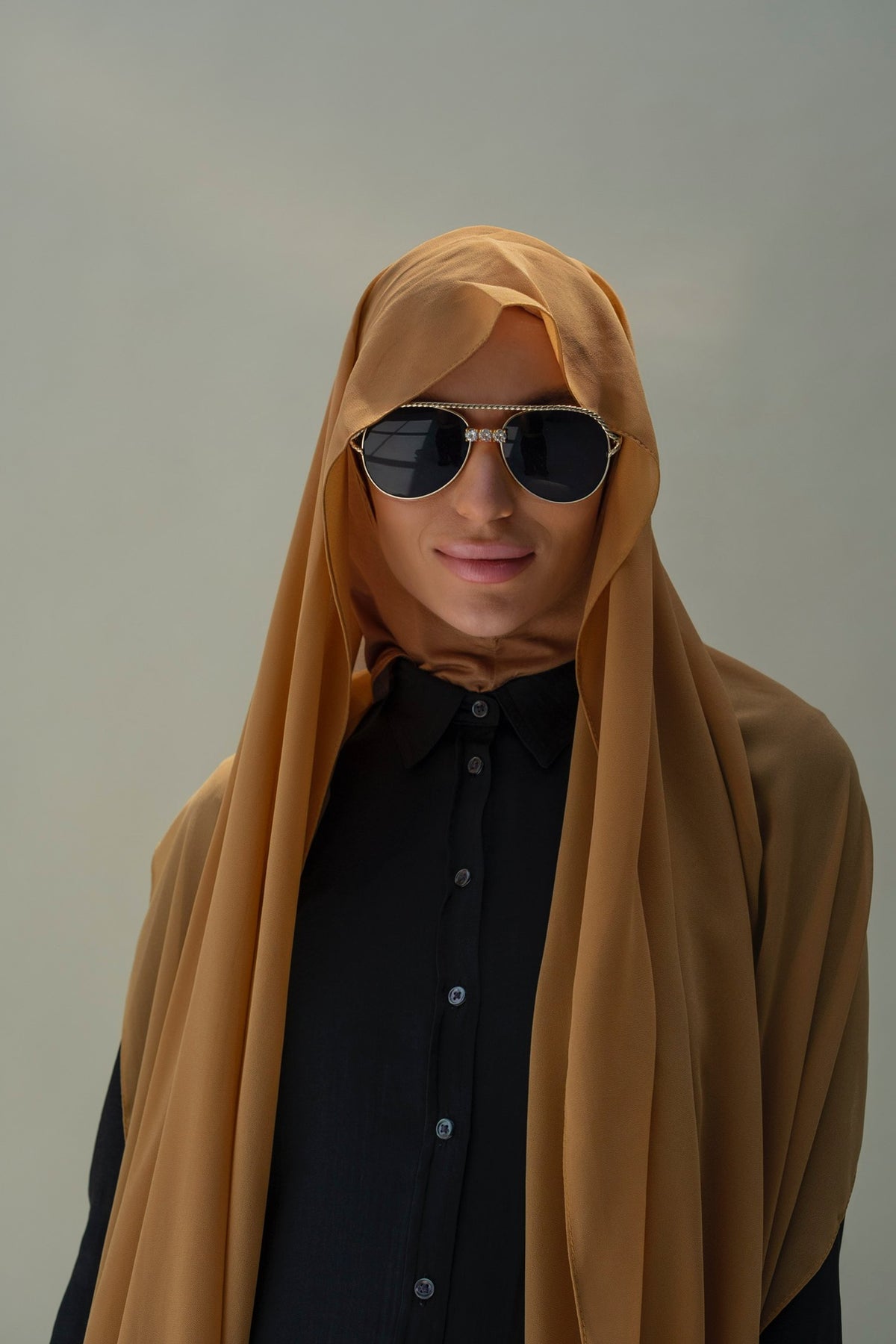 A person wearing a flowing mustard-yellow hijab and black sunglasses, layered over a black collared button-up shirt, set against a plain light gray studio background.