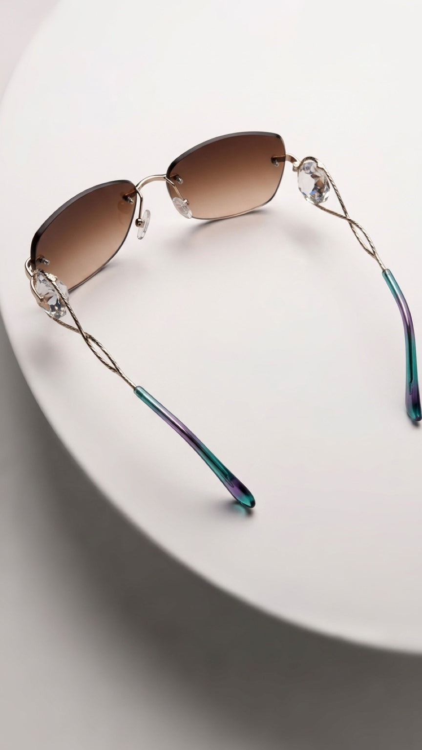 Rimless sunglasses with brown gradient lenses, clear rhinestone accents at the hinges, and iridescent green-blue temple arms, displayed on a white circular pedestal.