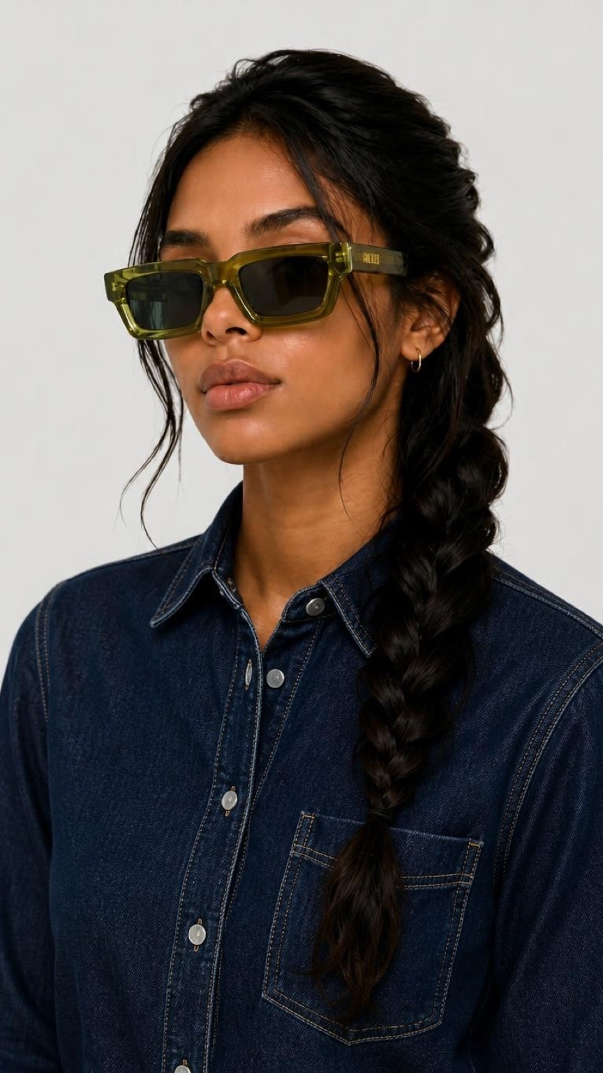 A woman with dark hair in a braid wearing olive green translucent square sunglasses and a dark blue denim shirt, photographed against a light gray background.