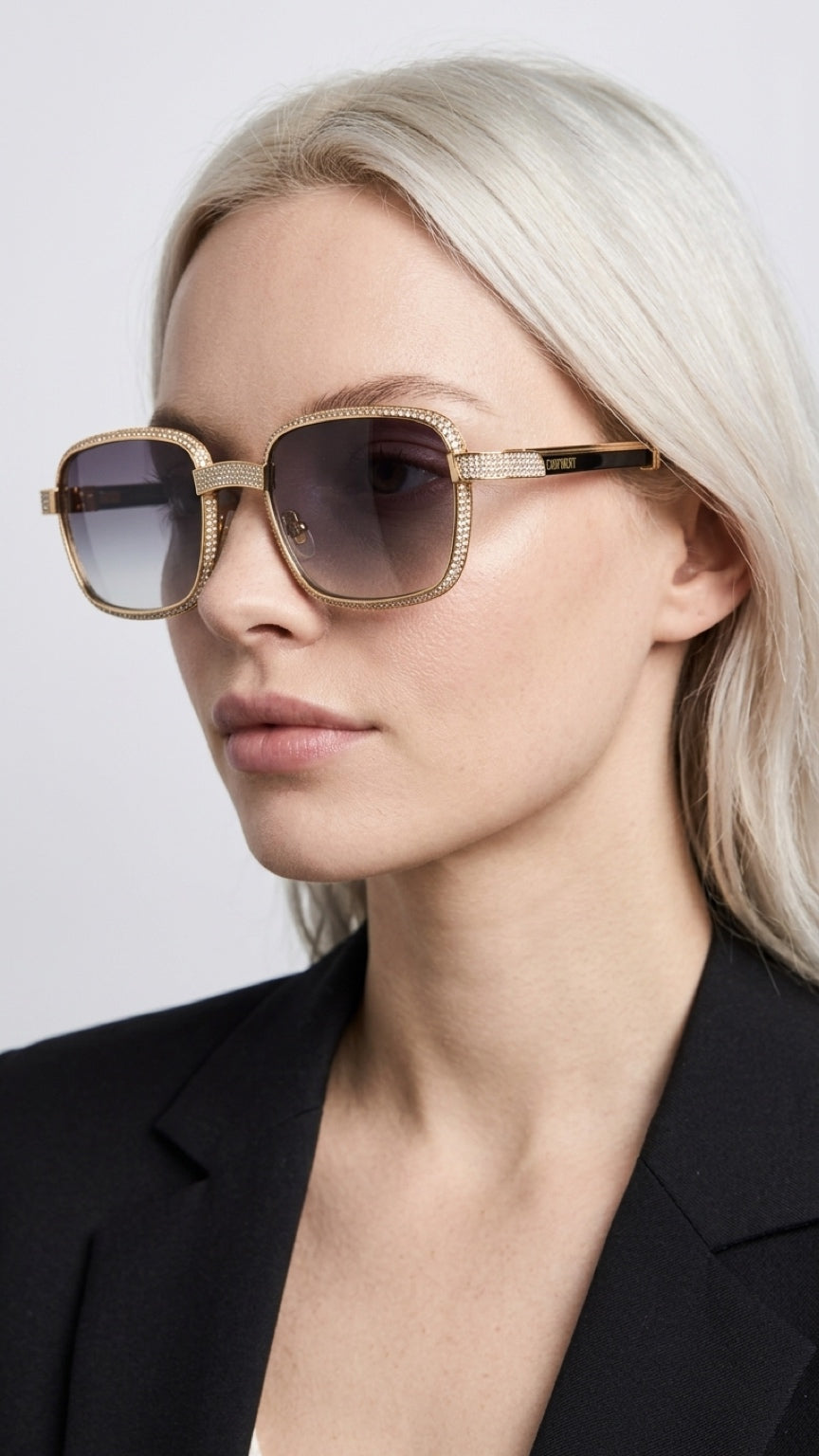 A close-up of a blonde woman wearing square sunglasses with a gold-tone metal frame and dark gradient lenses, paired with a black blazer against a light gray background.