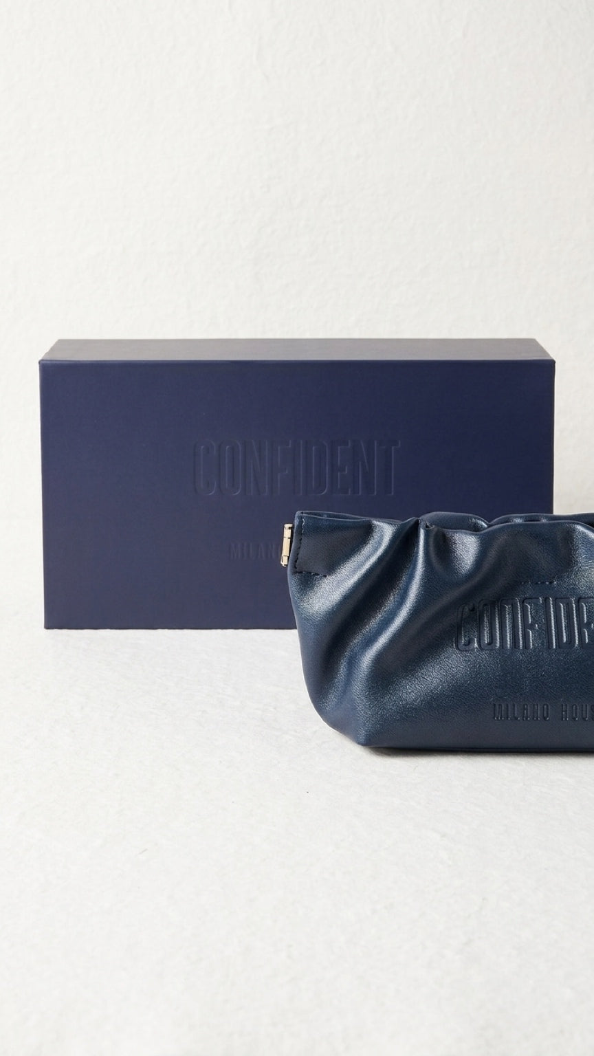 A navy blue leather pouch with a gold zipper and embossed Confident logo, shown in front of its matching navy blue presentation box on a light textured background.