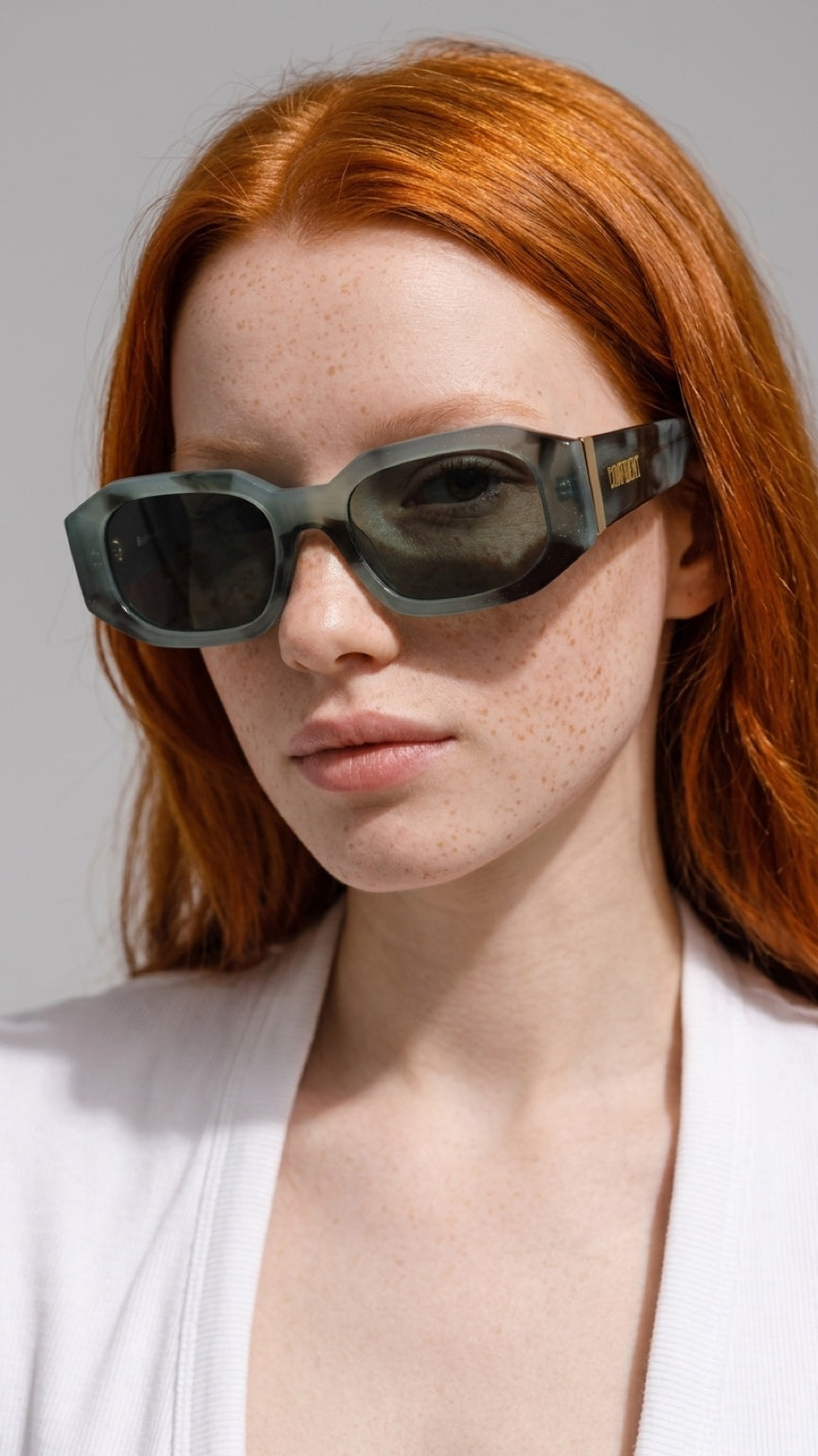 A close-up of a model with red hair wearing angular dark CONFILVARO Sage Green Marble-Effect Rectangle Sunglasses with a translucent gray-green frame and a white top against a light gray background.