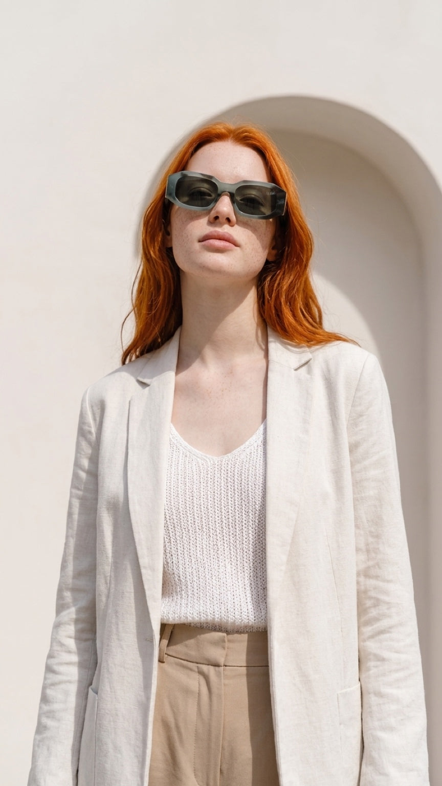 A woman with red hair wearing darkCONFILVARO Sage Green Marble-Effect Rectangle Sunglassessunglasses, a light beige blazer, a white knit top, and beige trousers, standing against a white stucco wall with an archway.