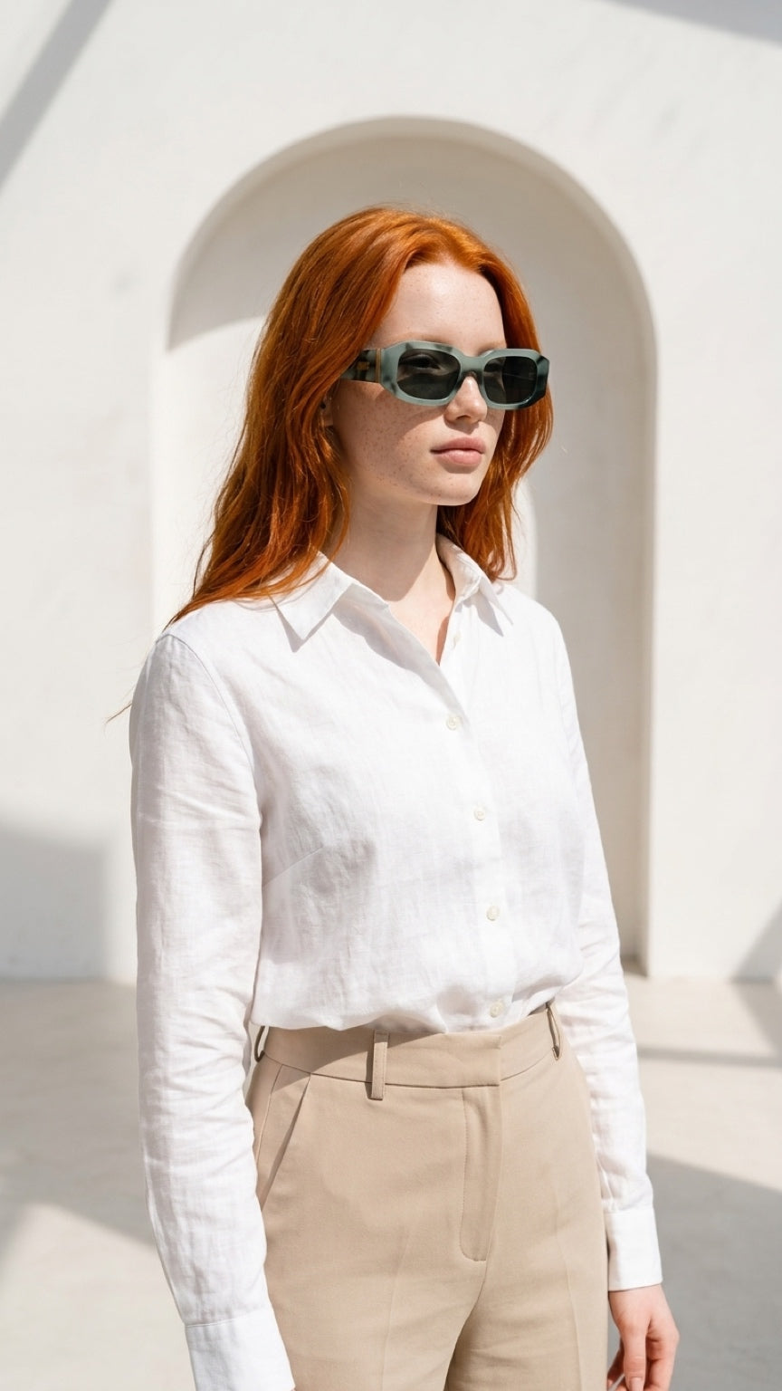 A woman with red hair wearing a white long-sleeve button-up shirt and beige trousers, styled with dark sunglasses, standing in front of a white arched wall.