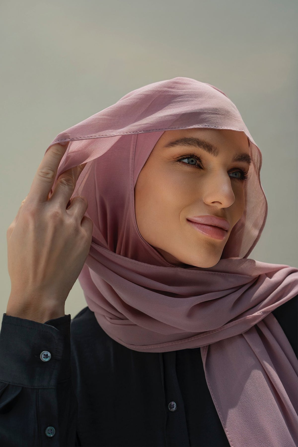 A woman wearing a dusty pink hijab and a black button-up shirt, posing against a soft, neutral gray background.