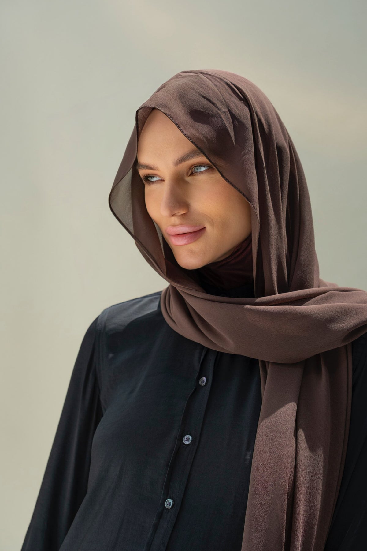 A woman wearing a draped brown hijab and a black button-up shirt, photographed against a soft light gray background.