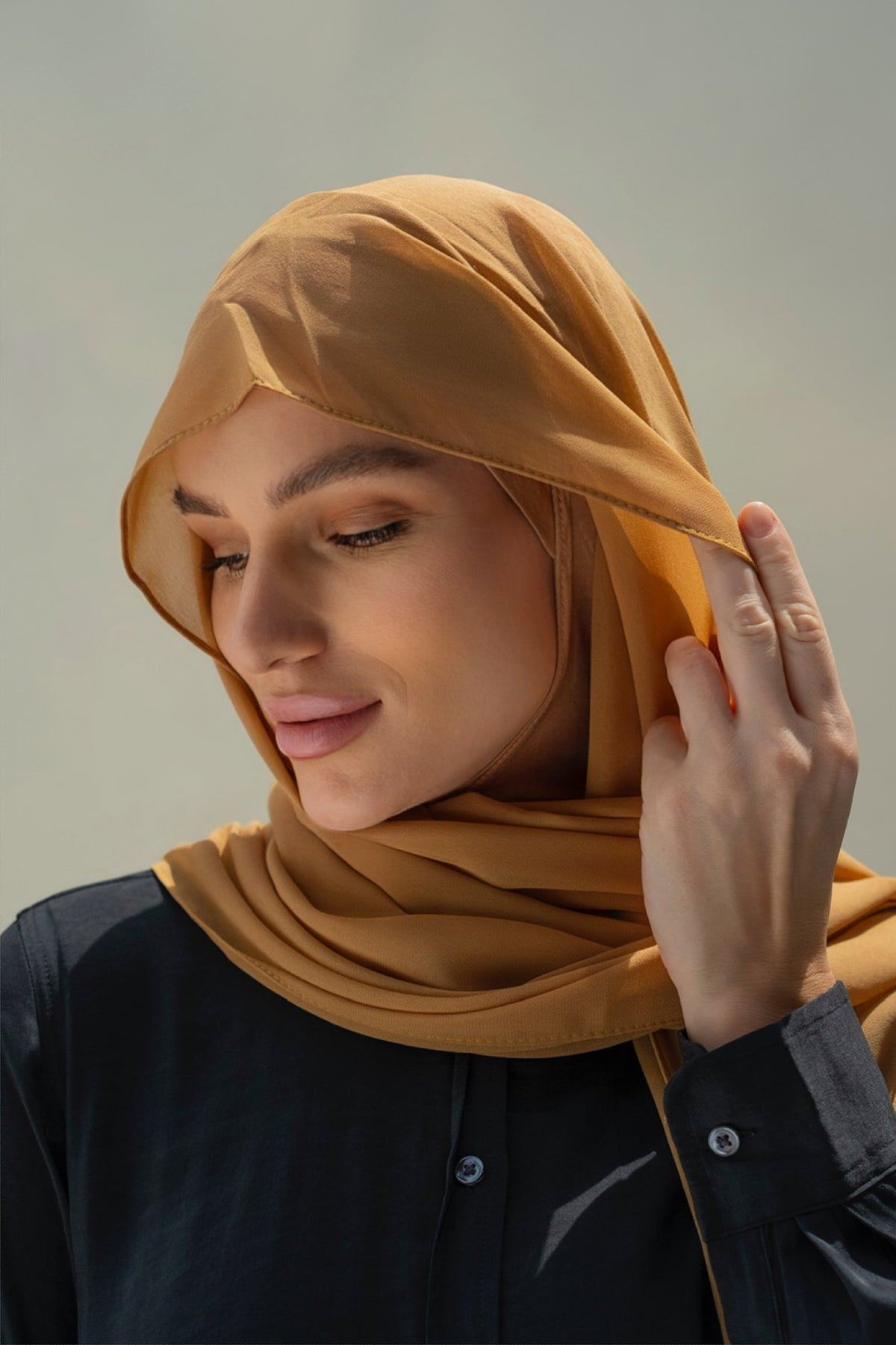 A woman wearing a mustard yellow hijab and a black button-up shirt, shown in a close-up portrait against a soft gray background.