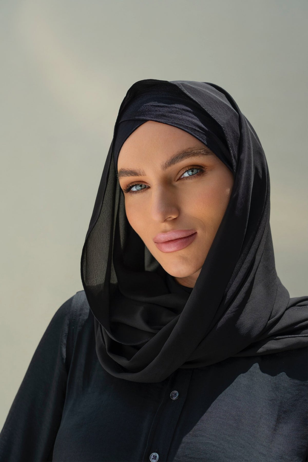 A close-up portrait of a woman wearing a black sheer hijab and a black button-front top against a soft neutral background.