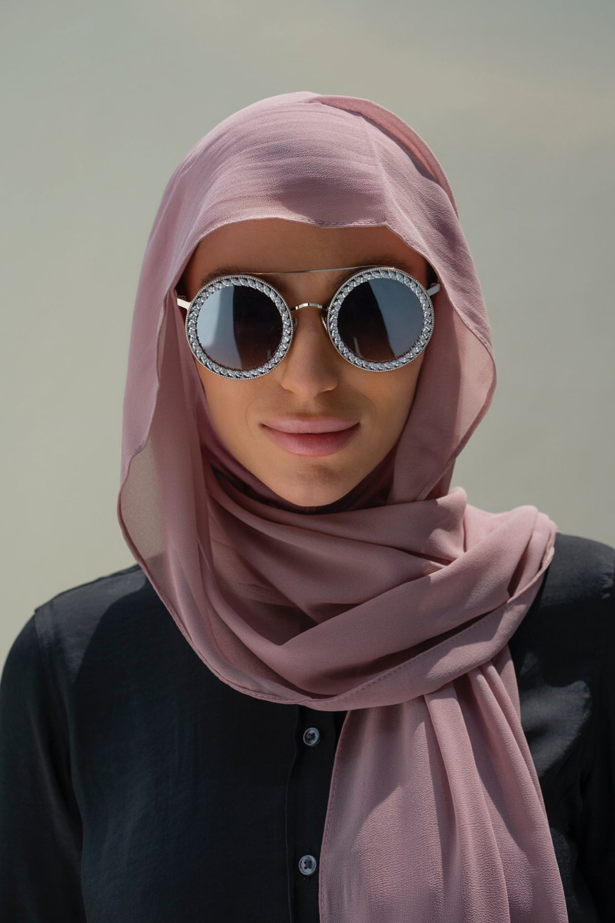 A woman wearing a dusty pink hijab and round silver-framed sunglasses with rhinestone trim, photographed against a soft neutral background.