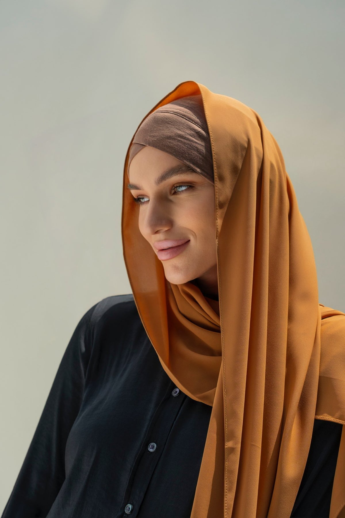 A woman wearing a brown scarf and inner cap, with a confident expression.