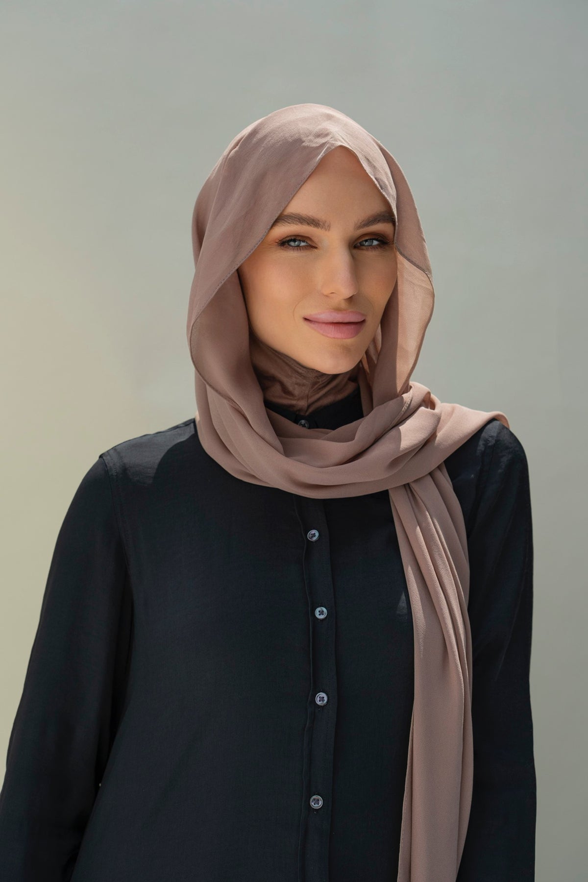 A woman wearing a dusty rose hijab and a black button-up top, photographed against a soft gray studio background.