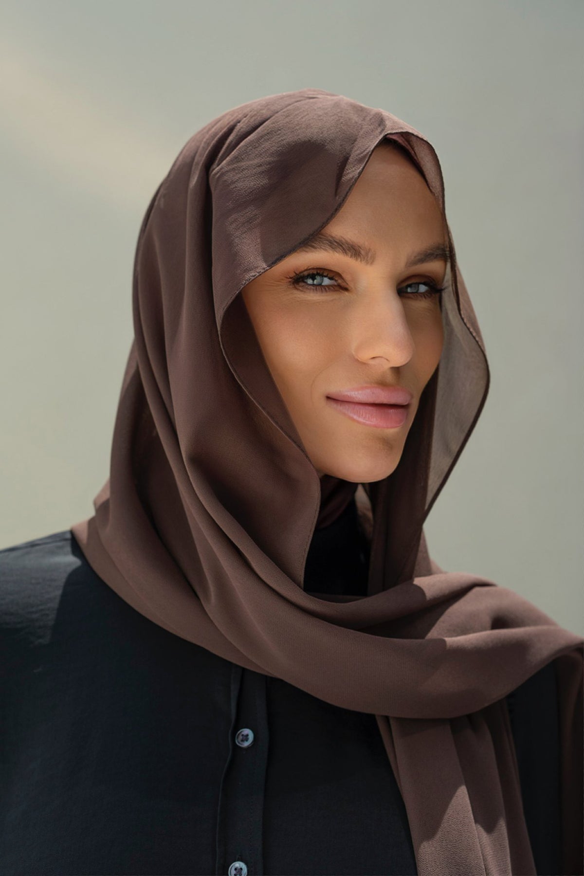 A close-up portrait of a woman wearing a draped taupe-brown hijab and a black button-up top against a soft neutral background.