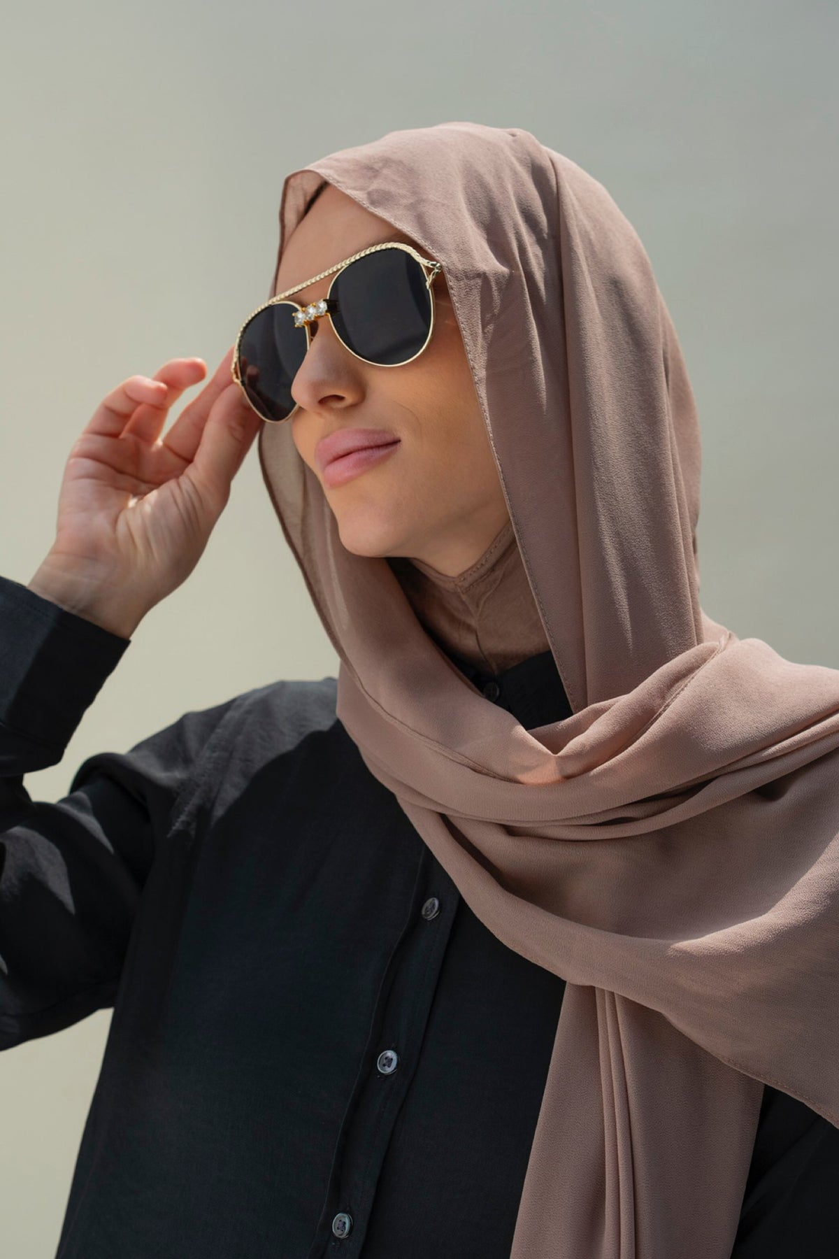 A woman wearing a taupe-brown hijab and gold-framed aviator sunglasses, dressed in a black button-up shirt against a plain light gray background.