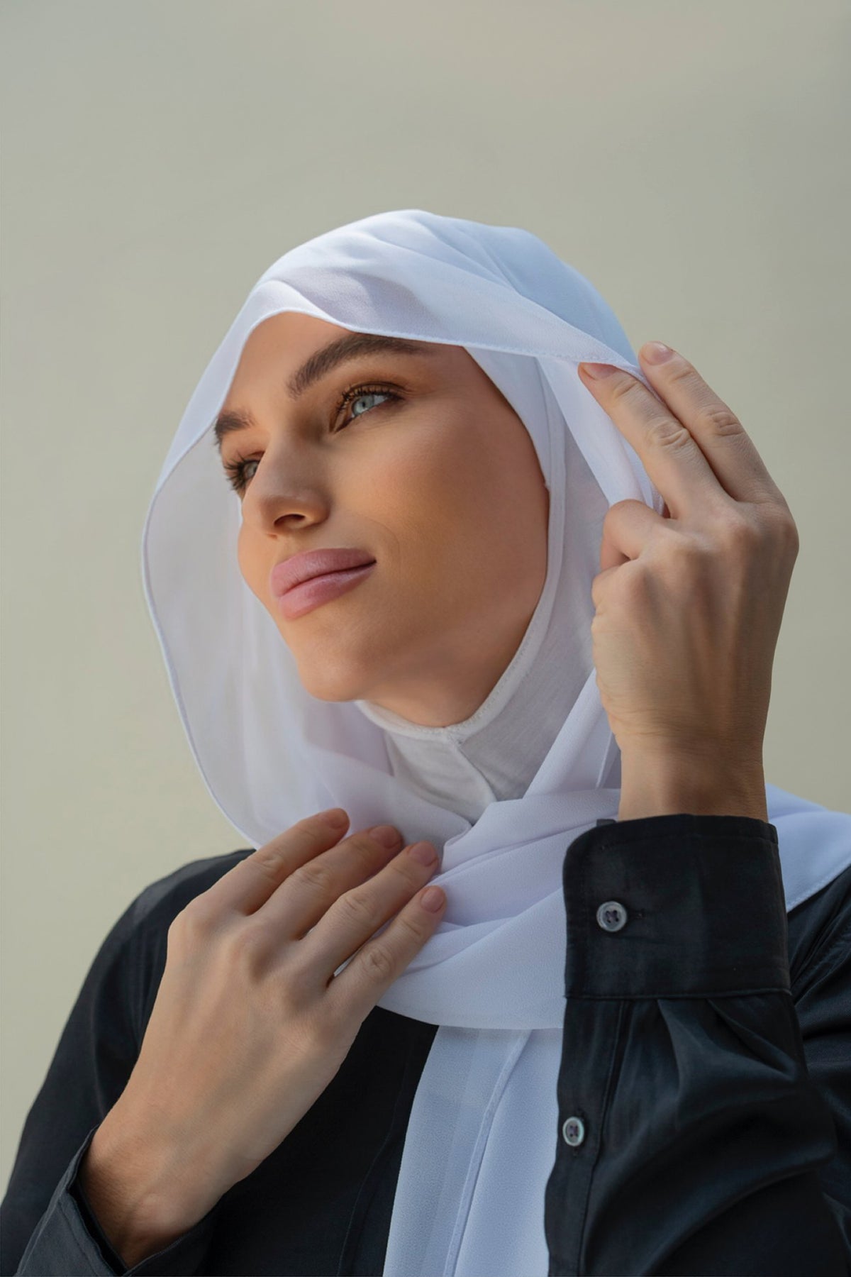 A woman wearing a white hijab and a black button-up shirt, posing against a neutral beige background.