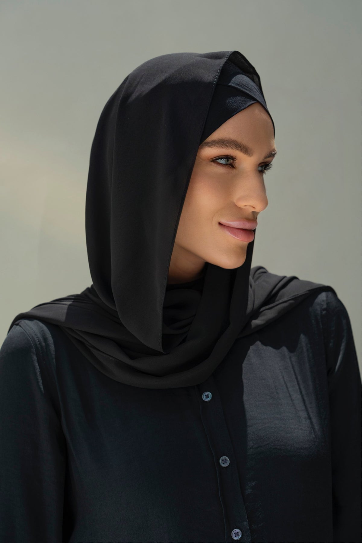 A woman wearing a black hijab and a black button-up top, shown in a close-up portrait against a soft light gray background.