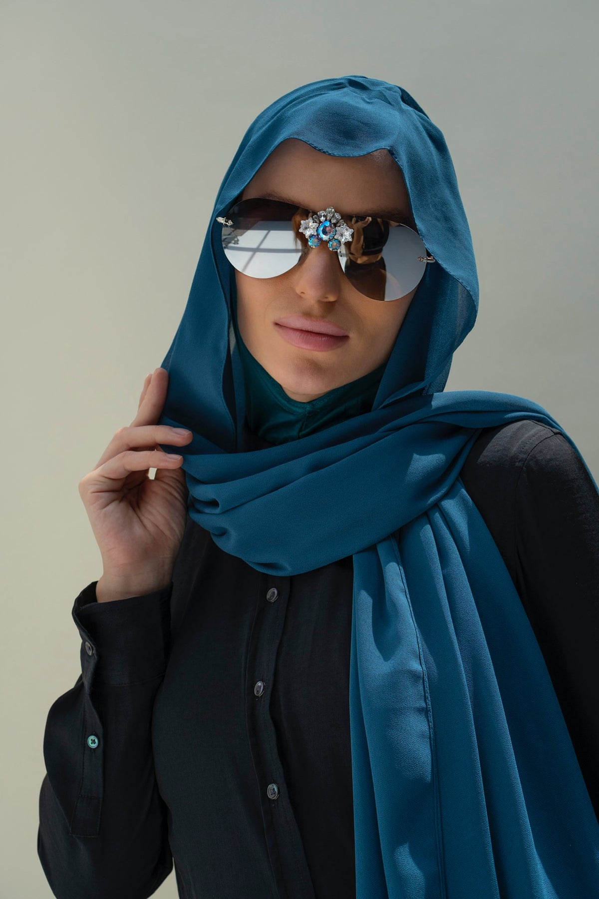 A woman wearing a teal blue hijab and a black button-up shirt, styled with decorative sunglasses, against a plain light gray background.
