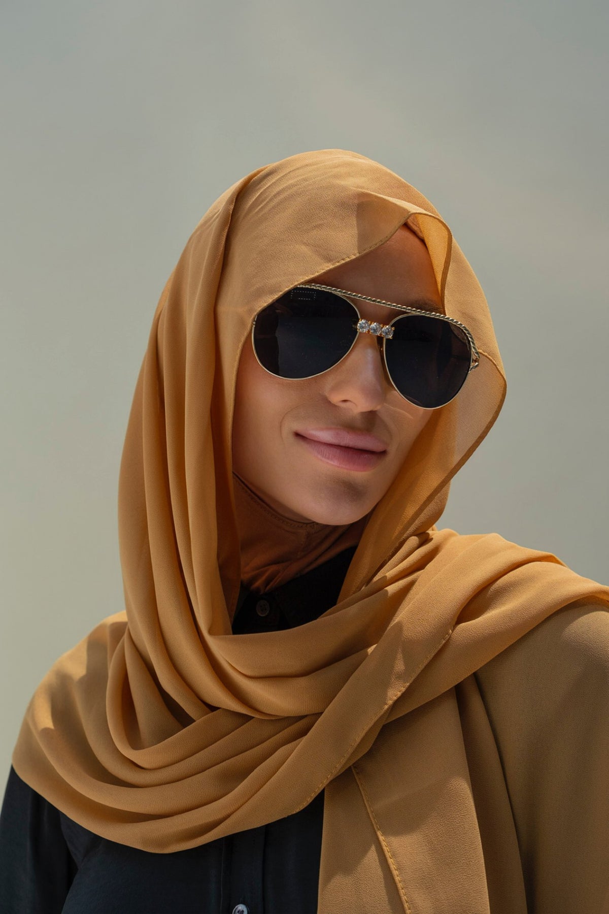 A woman wearing a mustard yellow hijab and dark sunglasses, shown in a close-up portrait against a soft gray background.