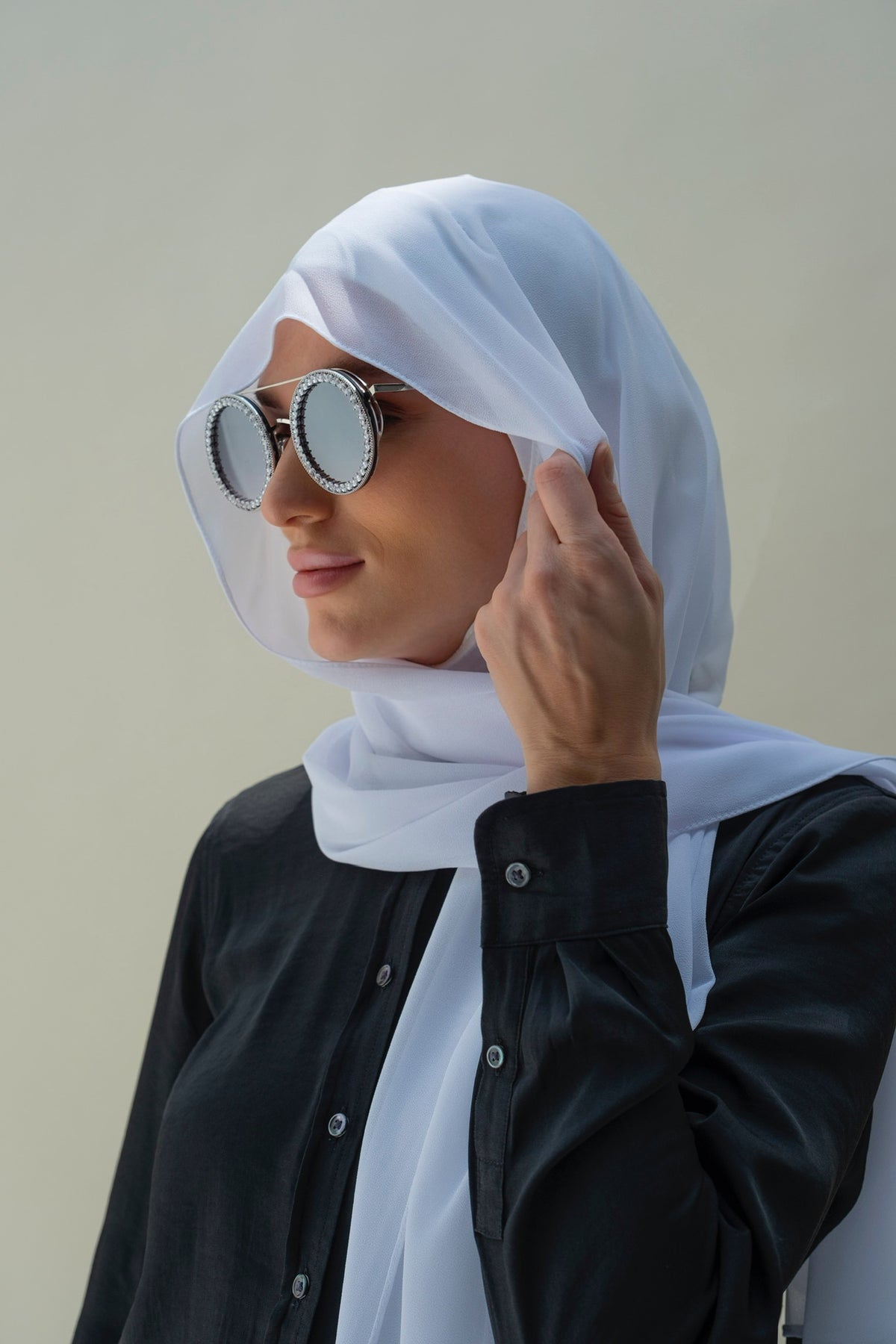 A woman wearing a white hijab and round silver-framed sunglasses, paired with a black button-up shirt, shown against a plain light beige background.