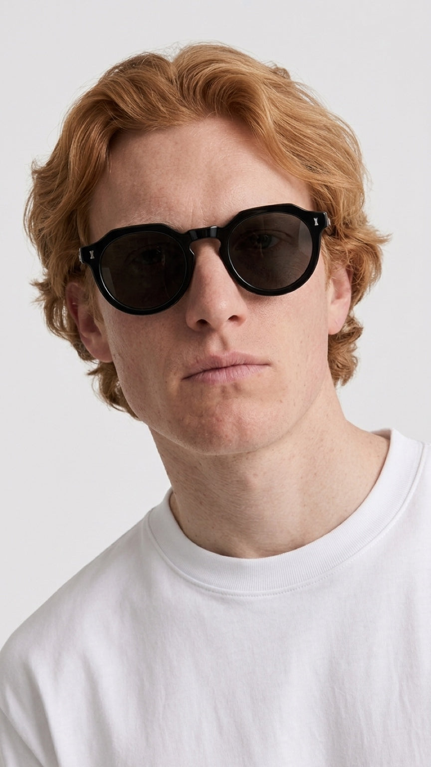 A close-up of a man with red hair wearing black round CONFILVARO Black Round Sunglasses
 and a white t-shirt against a light gray background.