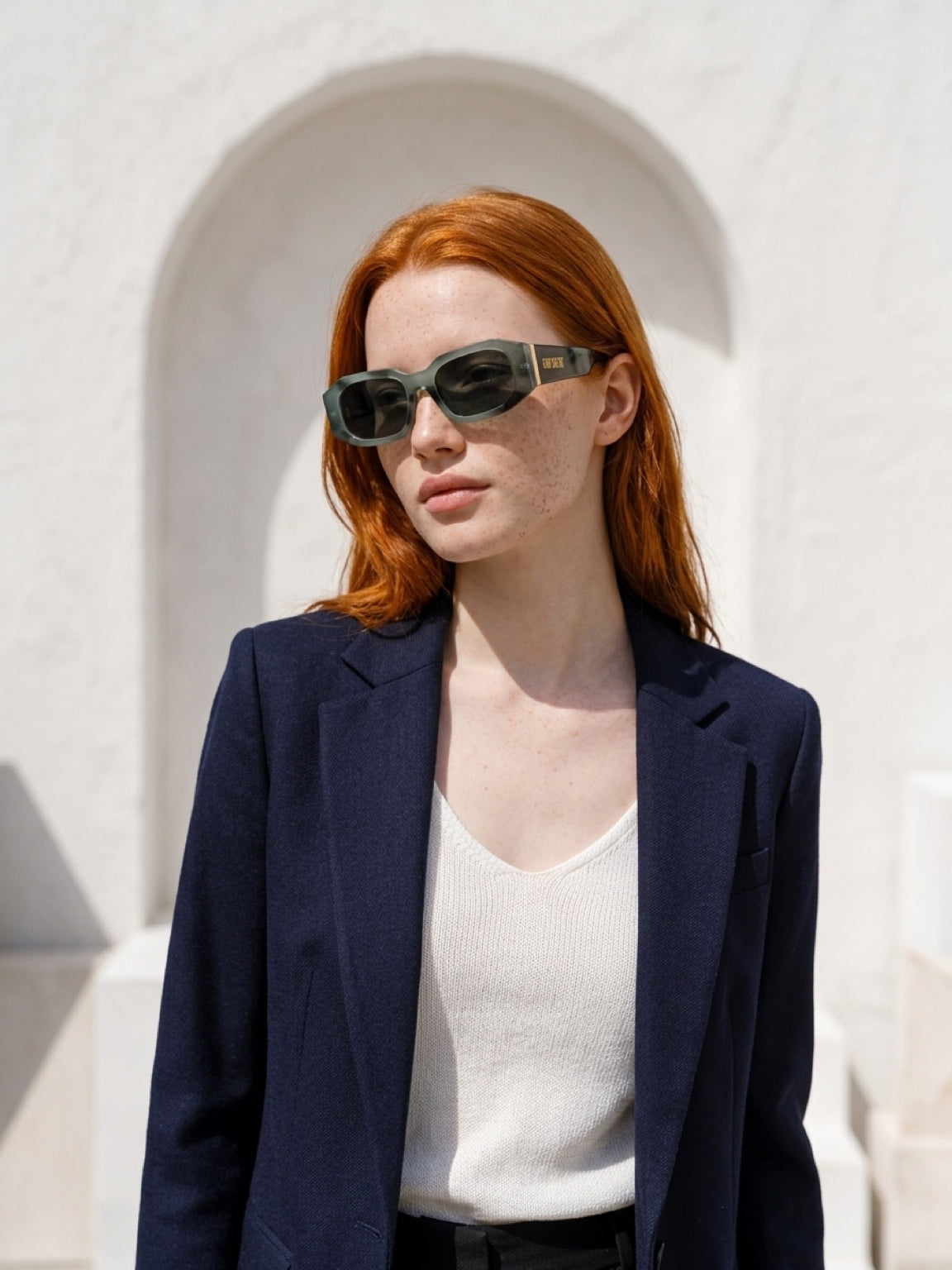 A woman with red hair wearing dark green-framed sunglasses, a navy blazer, and a white ribbed top, standing in front of a white stucco wall with an arch.