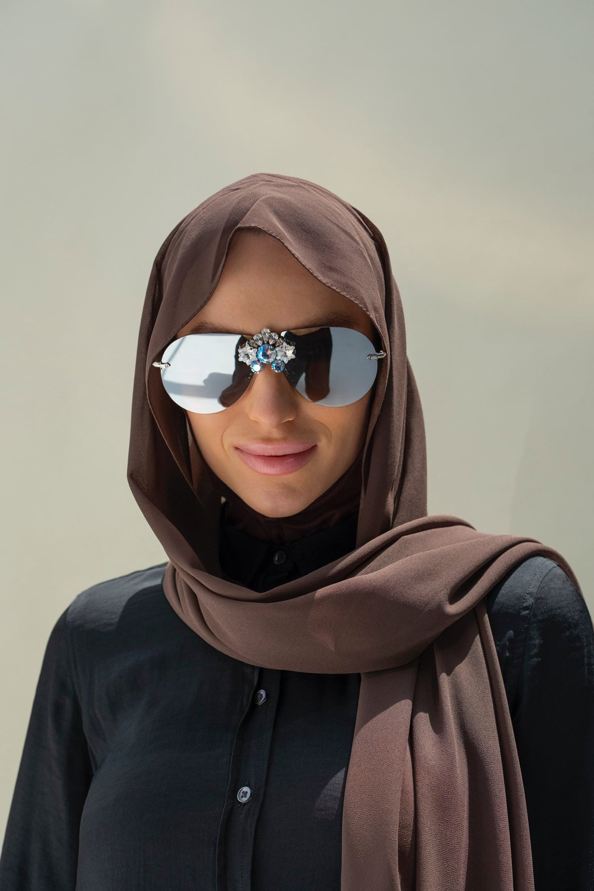 A woman wearing a brown hijab and dark sunglasses with a jeweled decorative bridge, set against a plain light gray background.