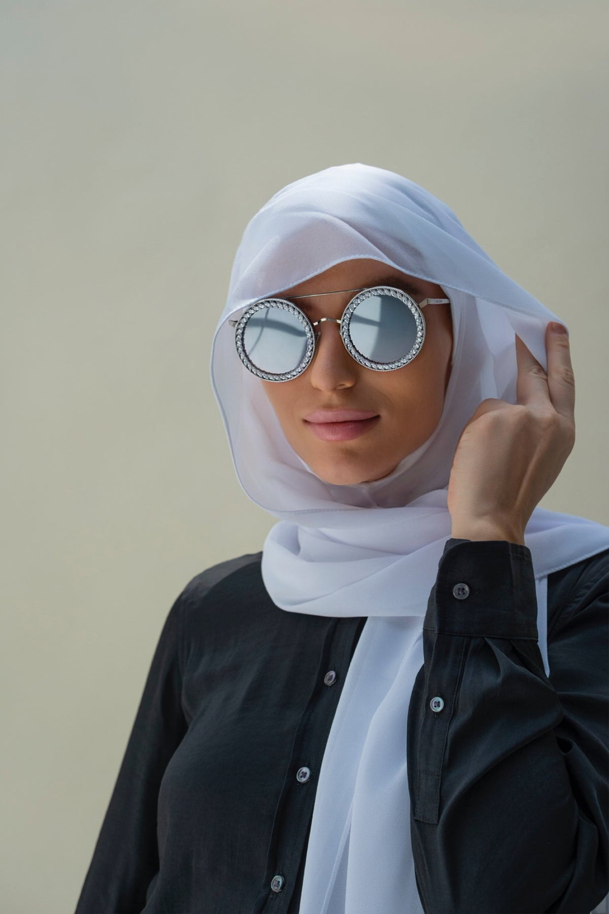 A woman wearing a white hijab and round silver-framed sunglasses with mirrored lenses, paired with a black button-up shirt, set against a plain beige background.