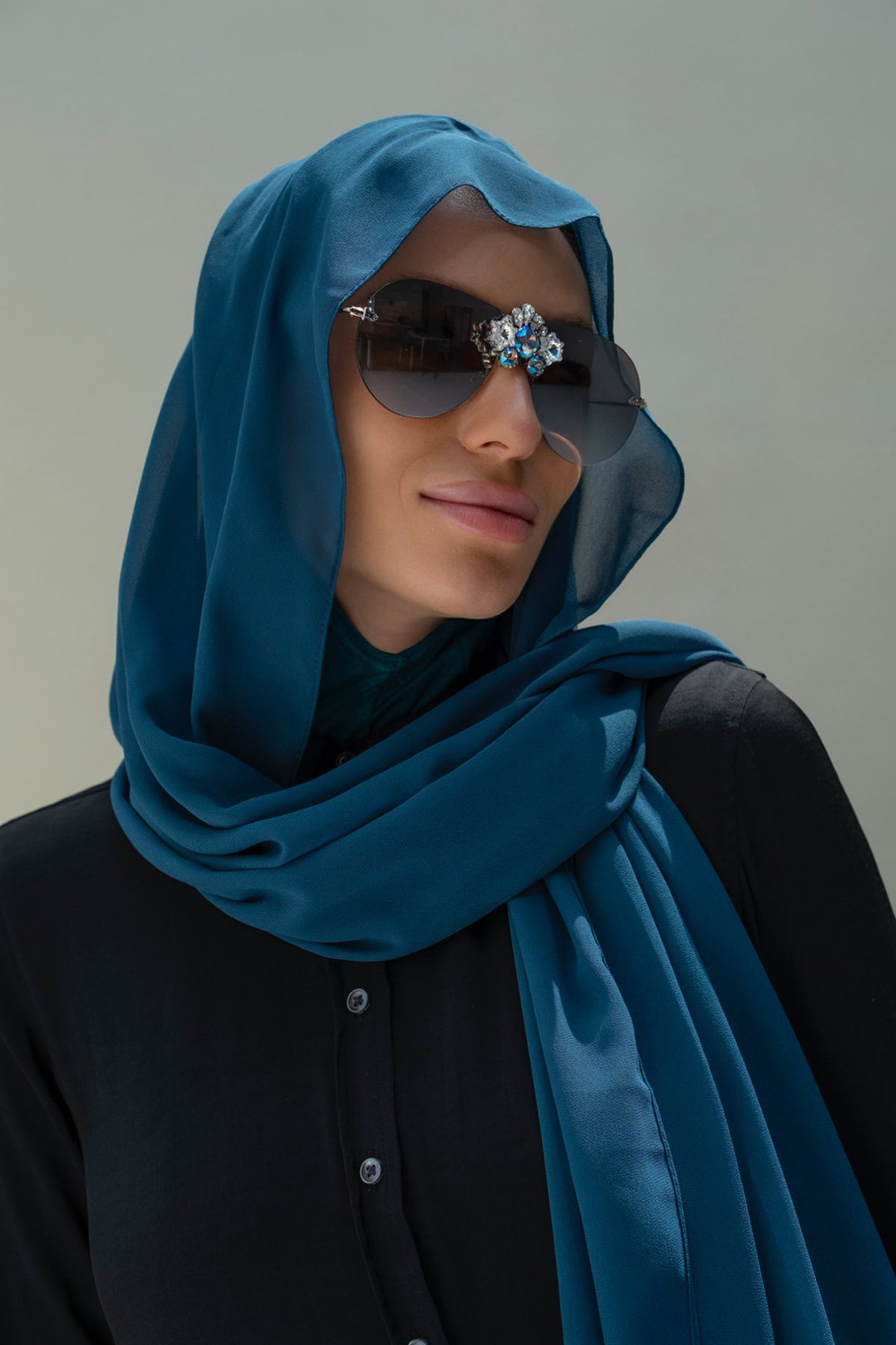 A woman wearing a teal blue satin hijab and black sunglasses with a jeweled bridge, dressed in a black top against a neutral gray background.