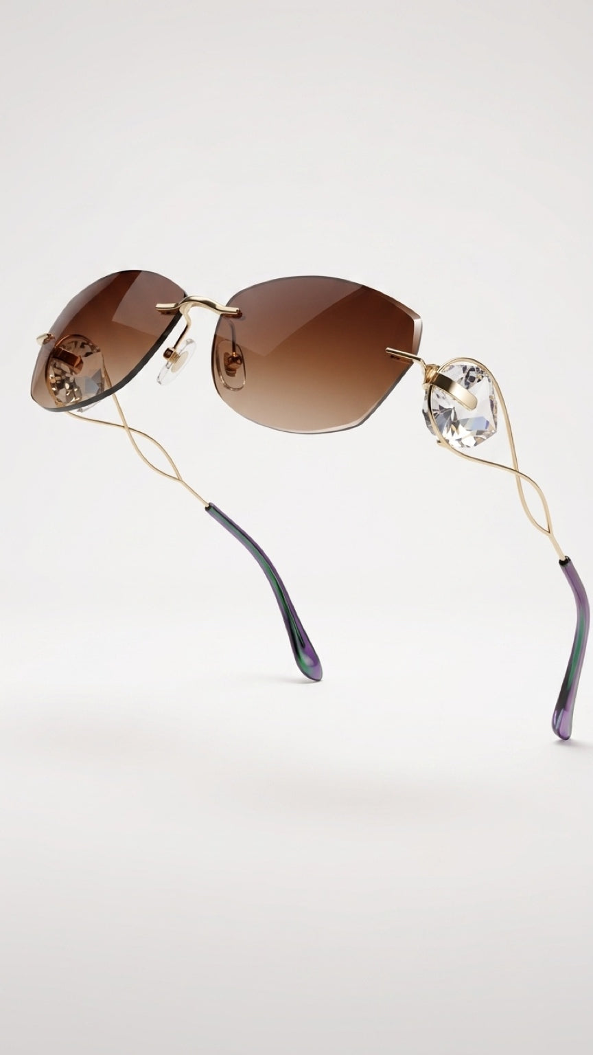 Rimless sunglasses with brown gradient lenses, a gold-tone metal frame, and clear rhinestone temples on a light gray background.