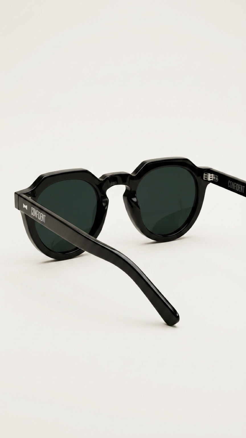 Black round sunglasses with dark green lenses and a silver-tone logo on the temple arm, photographed on a clean off-white background.