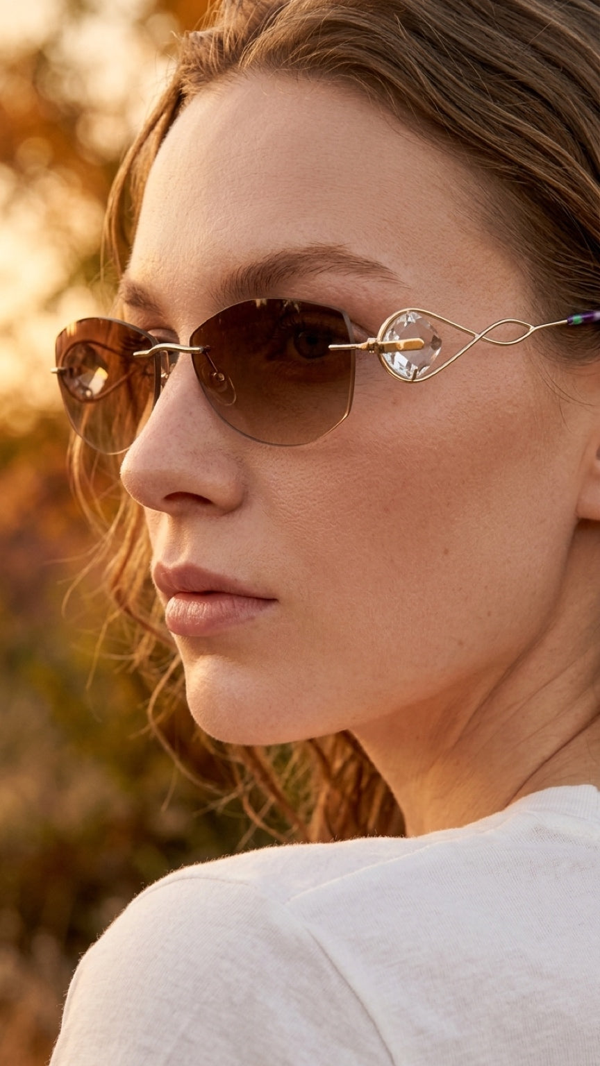 A close-up side profile of a woman wearing stylish oval sunglasses with thin gold metal frames, clear rhinestone accents, and brown tinted lenses, set against a warm, blurred outdoor background.