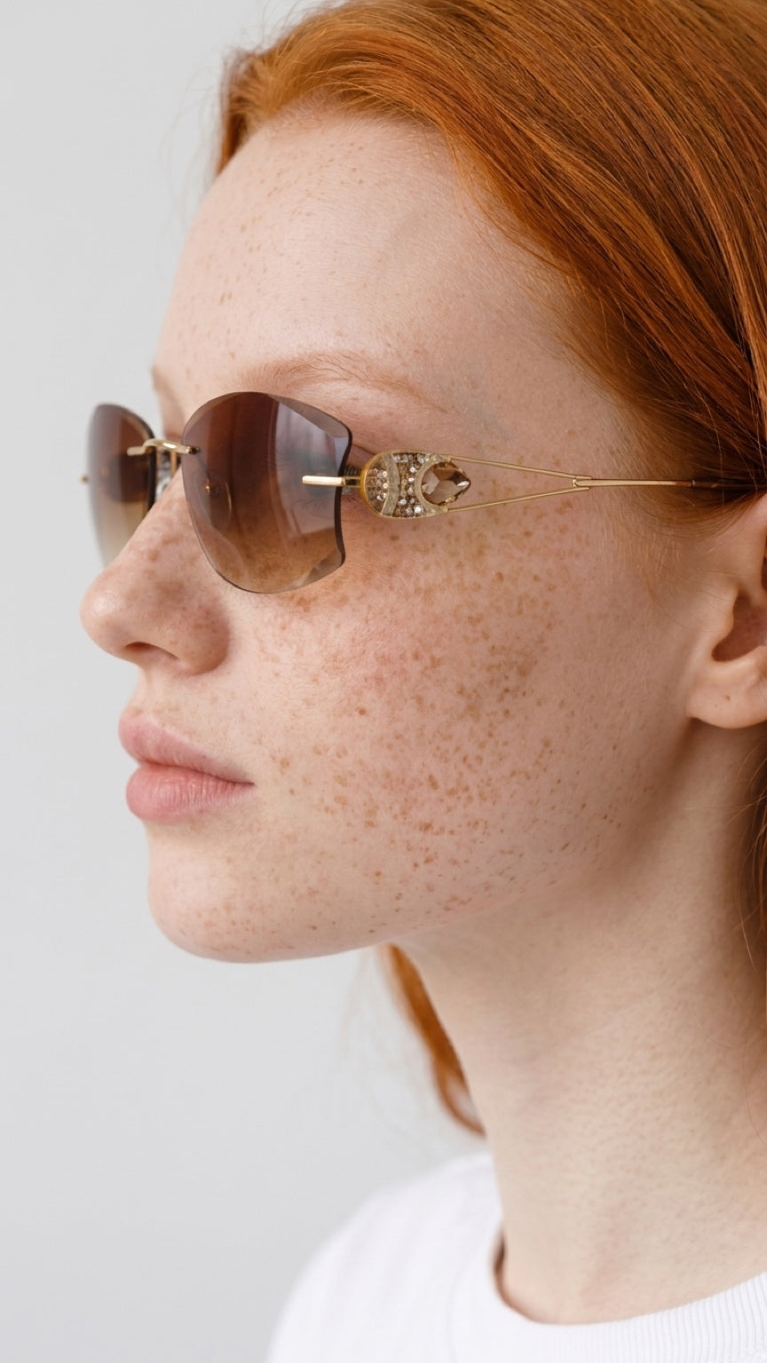 A close-up side profile of a woman with red hair and freckles wearing gold-framed sunglasses with a decorative crystal-embellished hinge and brown-tinted lenses, against a light gray background.