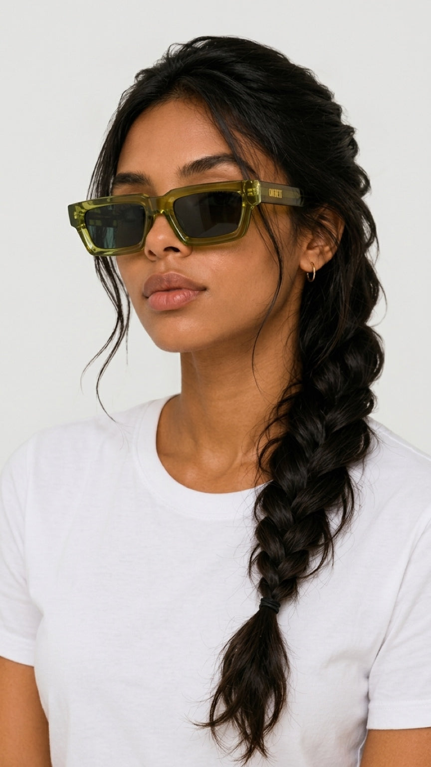 A woman with dark hair in a side braid wearing olive green translucent square sunglasses and a white t-shirt, photographed against a light gray background.