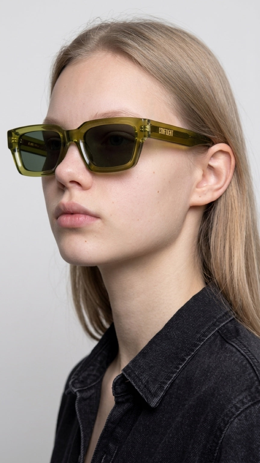 A close-up side profile of a model wearing olive green translucent square sunglasses with dark lenses and a black denim shirt against a light gray background.