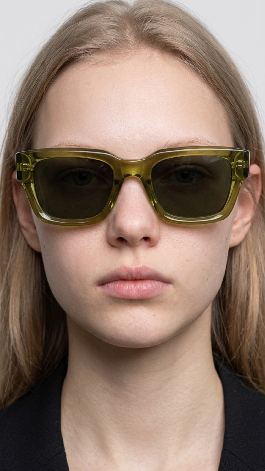 A close-up of a model wearing oversized square sunglasses with translucent olive green frames and dark lenses.