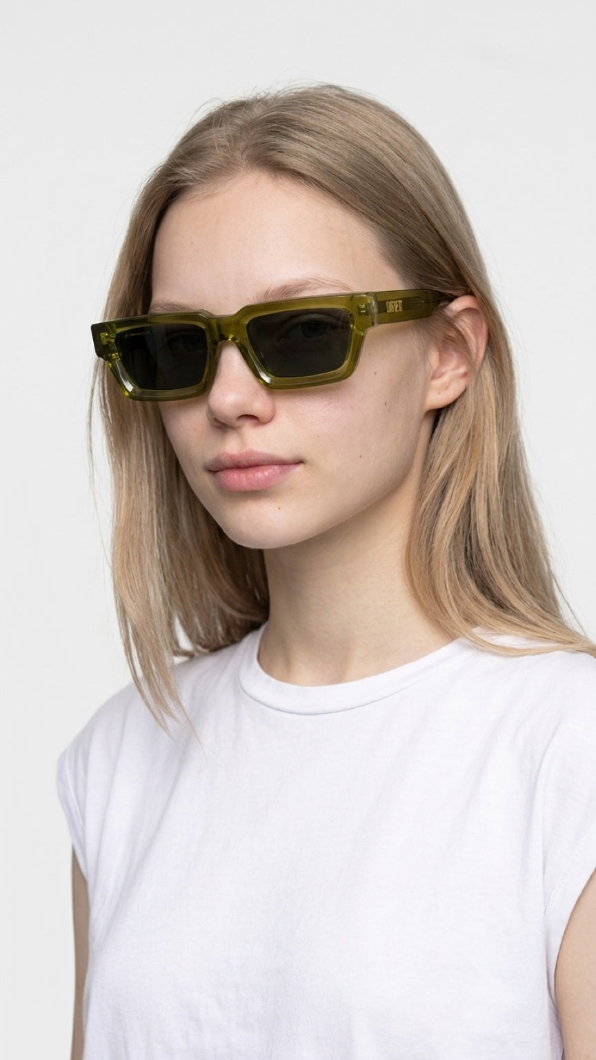 A woman with blonde hair wearing translucent olive green square sunglasses and a white t-shirt, photographed against a clean white background.