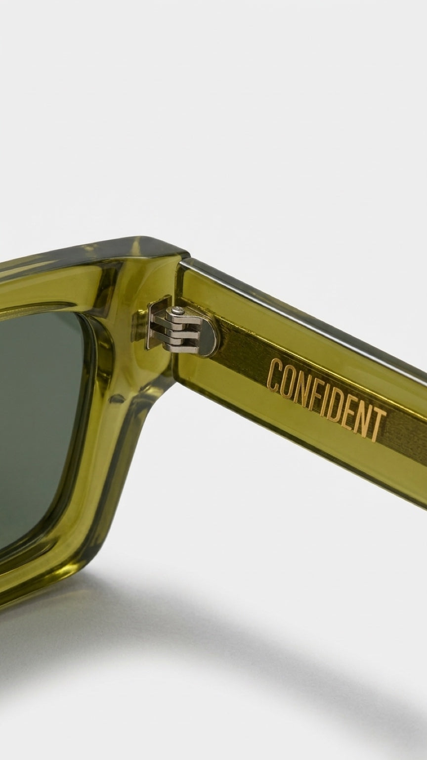 Close-up of olive green acetate sunglasses with dark green lenses and gold 'CONFIDENT' branding on the temple arm, set against a light gray background.
