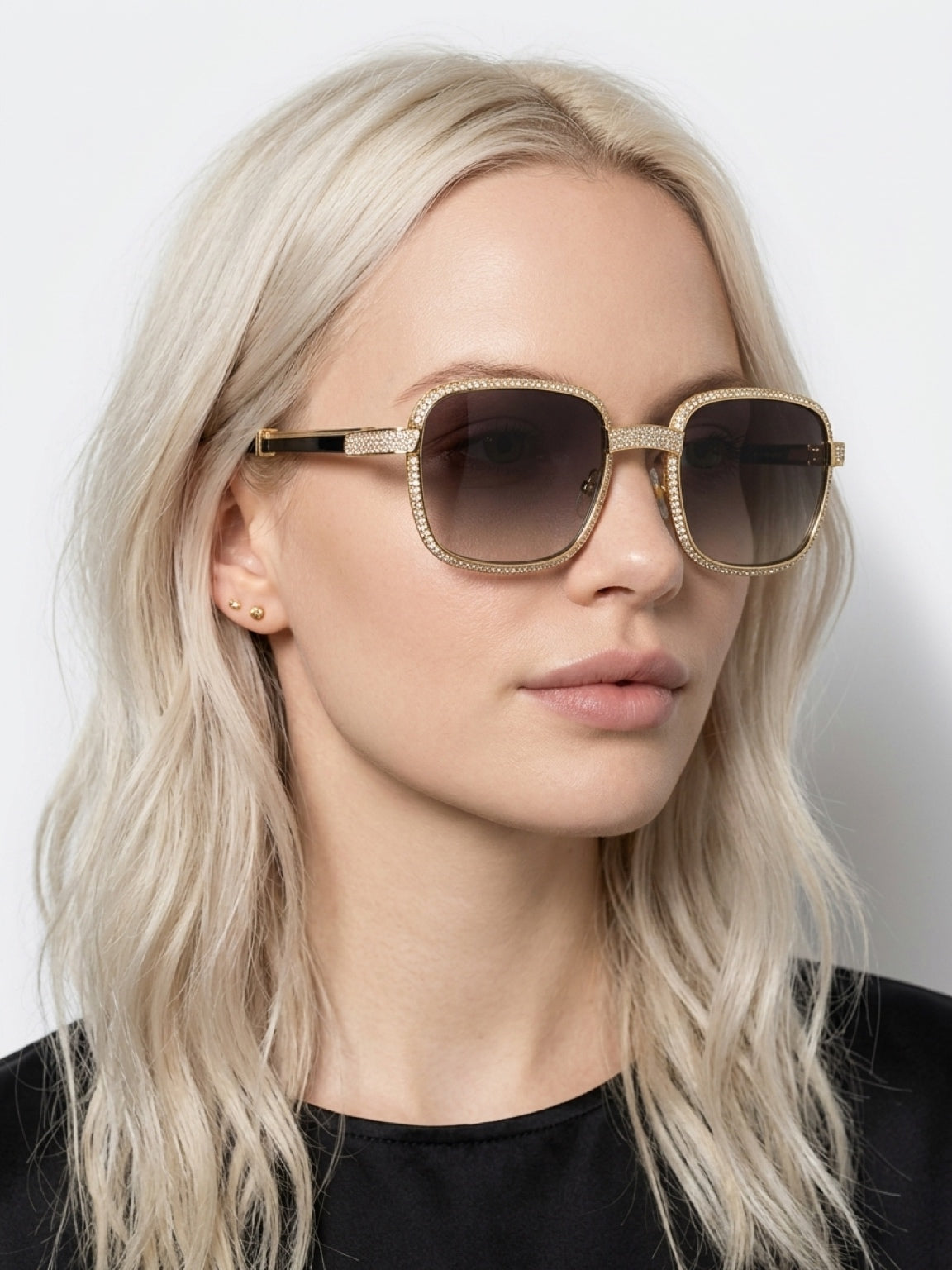 A close-up of a blonde woman wearing oversized square sunglasses with a gold-tone frame embellished with small clear rhinestones and dark tinted lenses.